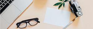 Top-down view of a minimalist workspace featuring a blank white card, black-rimmed glasses, and green leaves on a light wood surface.